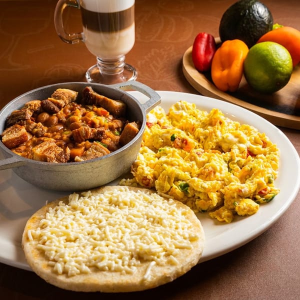 Breakfast plate with stew, scrambled eggs, cheesy flatbread, a latte, and fresh produce.