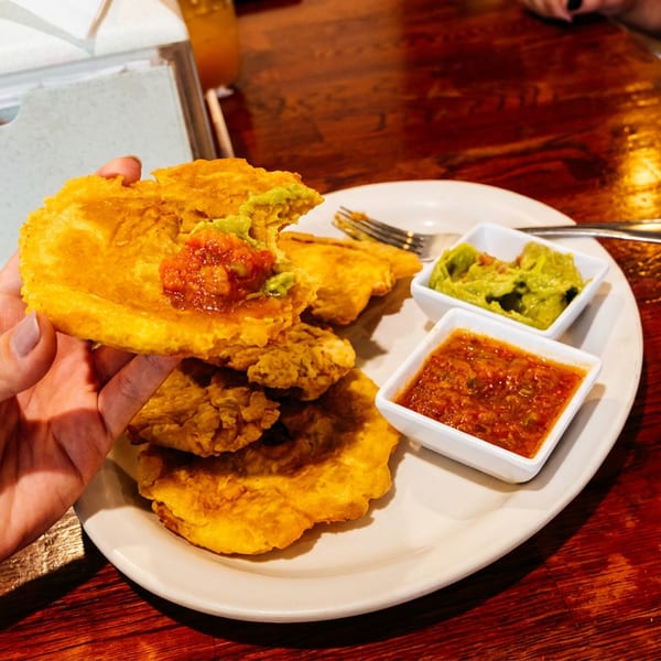 Hand holding a fried plantain topped with salsa and guacamole, with more on a plate.