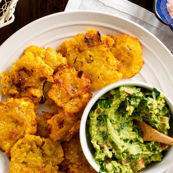 Crispy fried plantains and a bowl of chunky guacamole with a wooden spoon on a white plate.