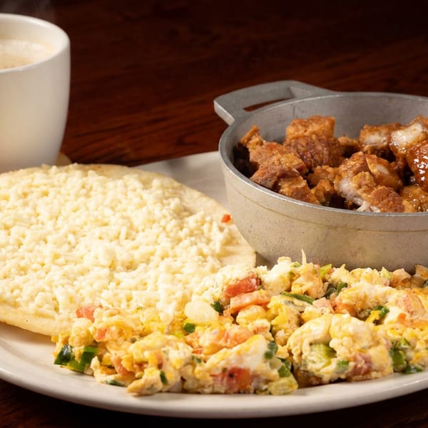 Breakfast plate with cheese arepa, scrambled eggs, chicharrones, and a steaming coffee cup.
